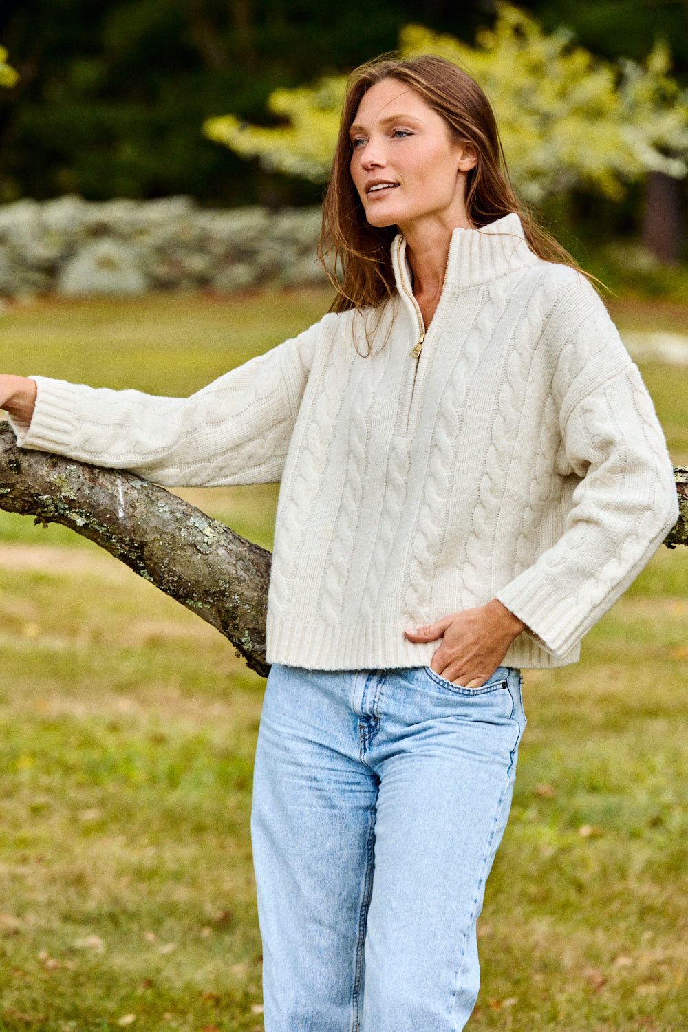 Woman wearing a white cable knit sweater and light blue jeans standing outdoors.