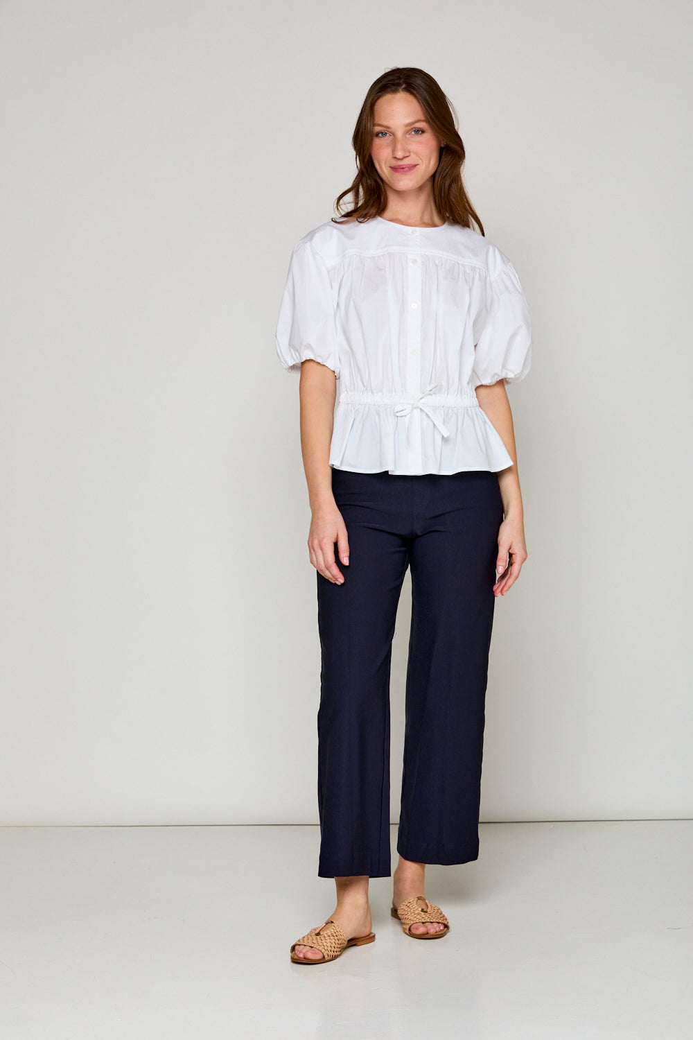 A woman wearing a white Laguna Blouse with a jewel neckline and puff sleeves, paired with navy pants.