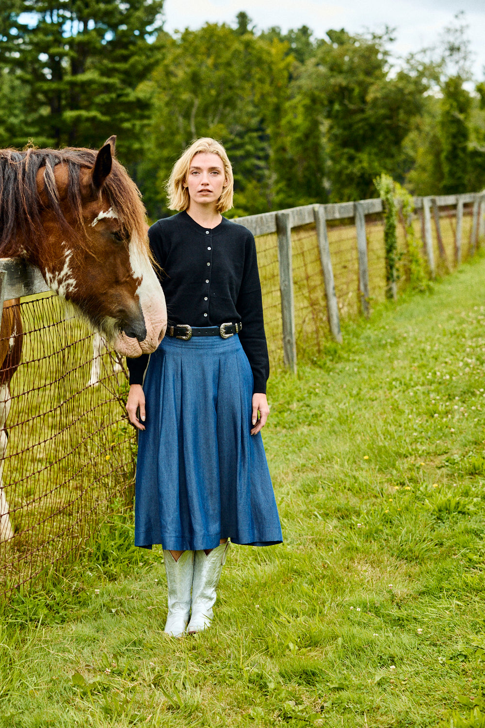 Woman in a blue skirt standing next to a horse in a grassy field with a wooden fence and trees in the background.