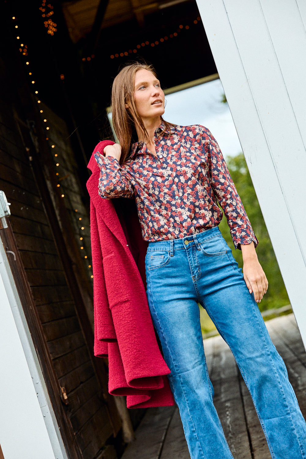 Woman in a patterned shirt and blue jeans holding a red coat, standing on a wooden porch.