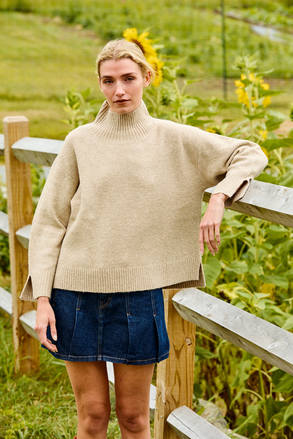 Woman wearing a beige sweater and denim skirt standing in a garden with sunflowers.