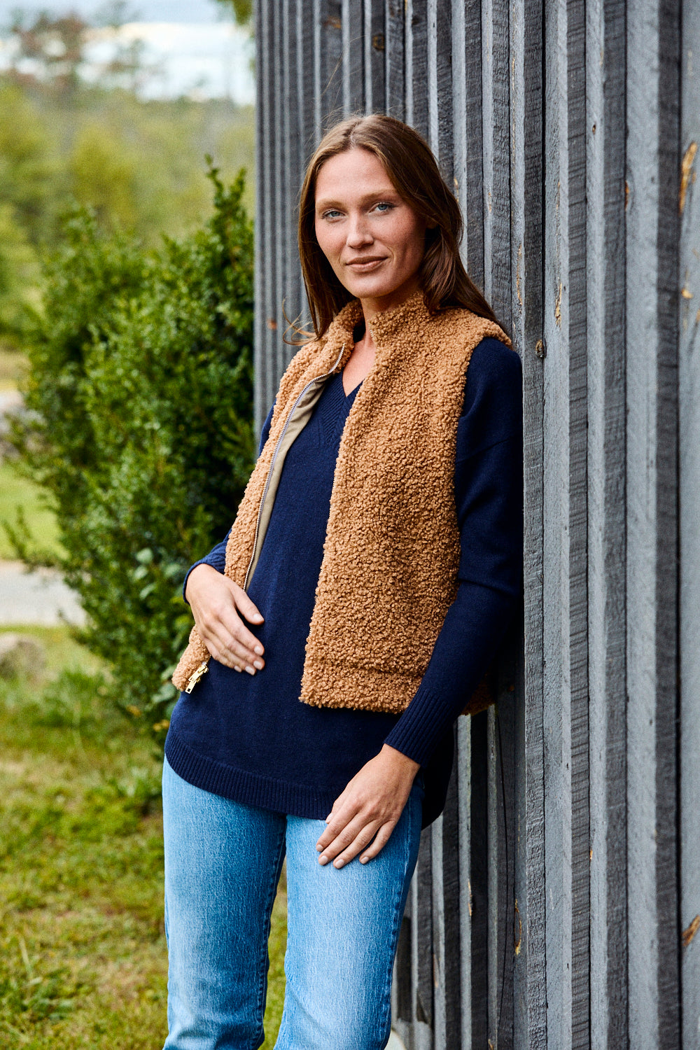 Woman wearing a brown vest over a navy sweater and blue jeans, standing against a corrugated metal wall.