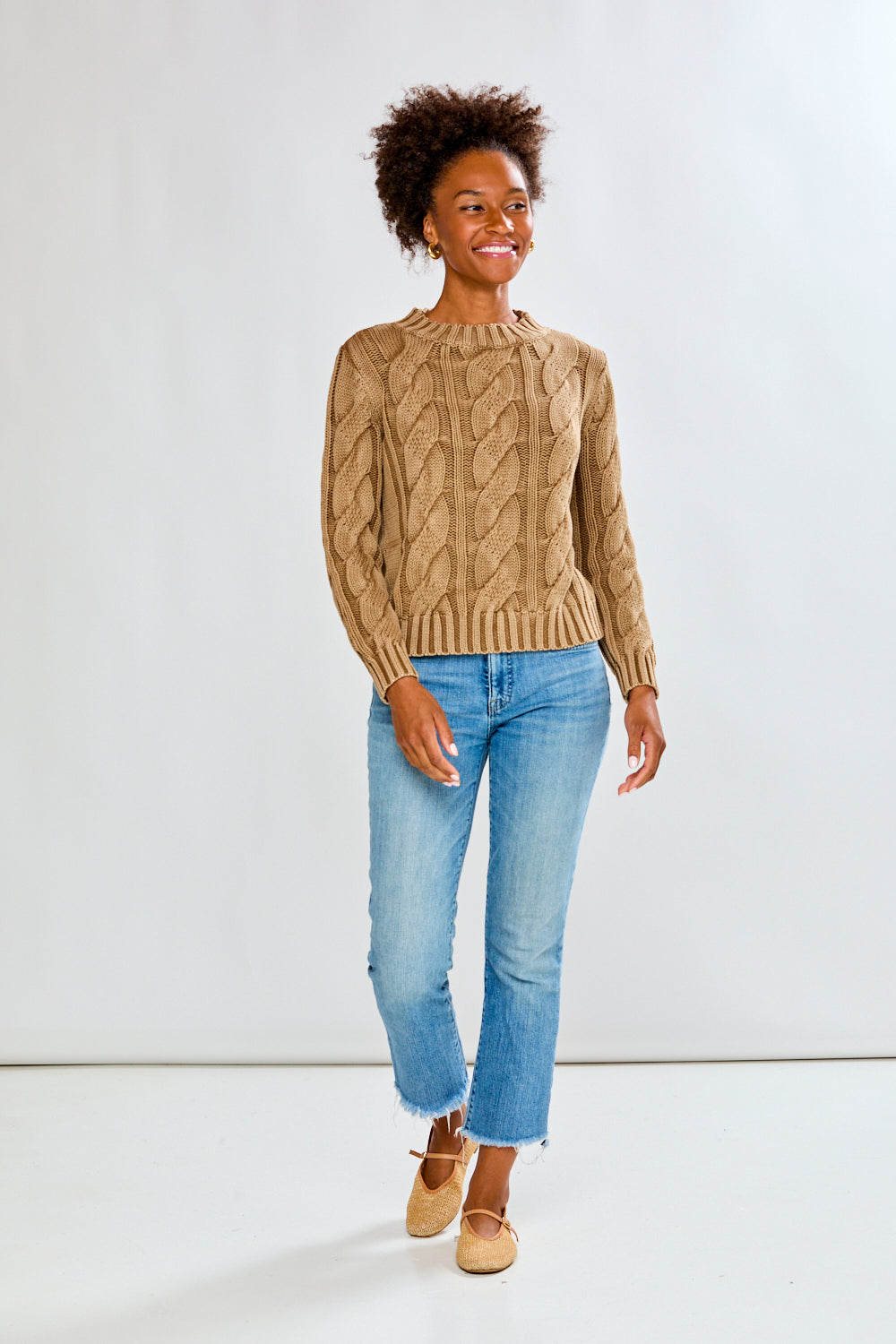 Woman wearing a toffee cable knit sweater and blue jeans on a white background