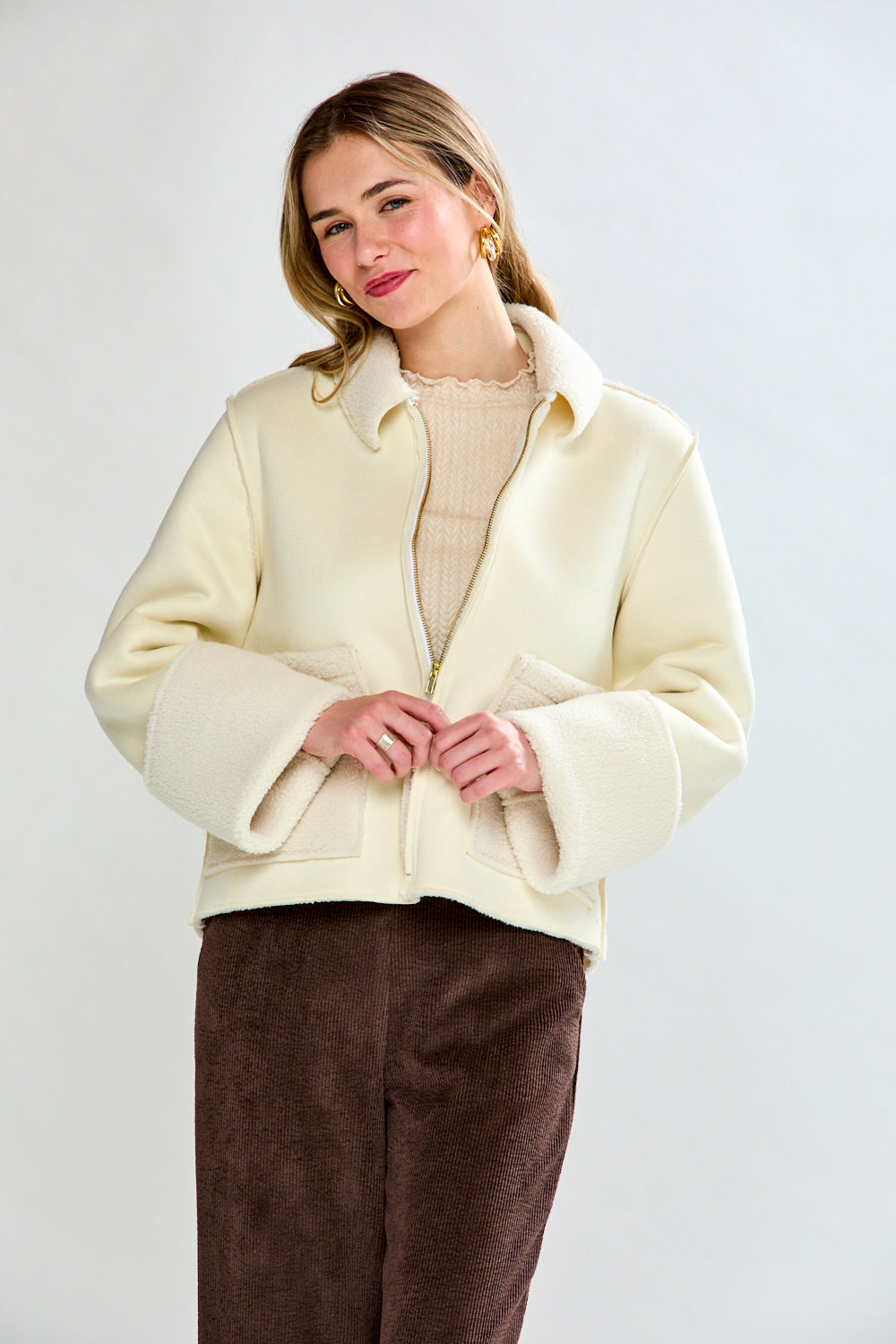 Woman wearing a cream-colored coat over a beige sweater and brown pants on a white background
