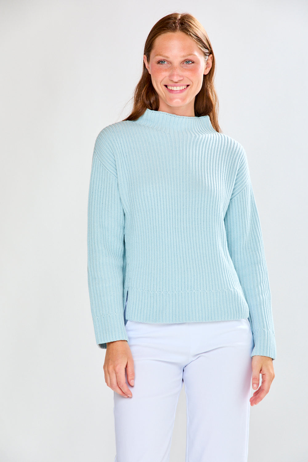 Woman wearing a light blue ribbed sweater and white pants on a plain background