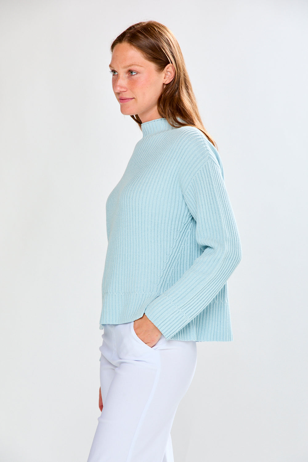 Woman wearing a light blue ribbed sweater and white pants on a plain background