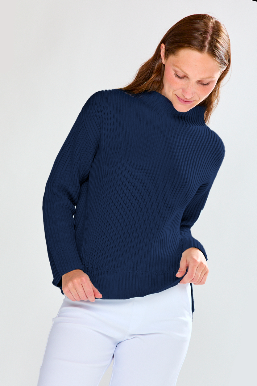 Woman wearing a navy ribbed sweater and white pants on a plain background