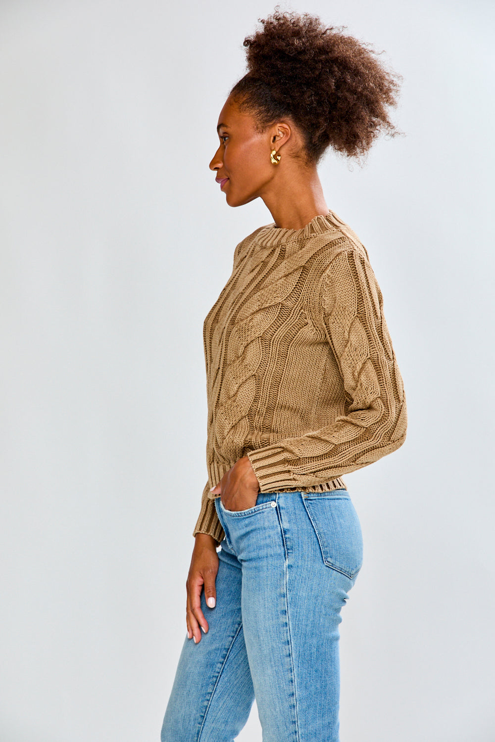 Woman wearing a toffee cable knit sweater and blue jeans on a white background