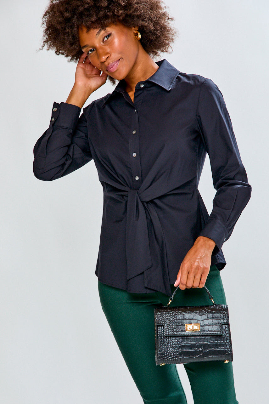 Woman wearing a black shirt and green pants, holding a black handbag against a light gray background