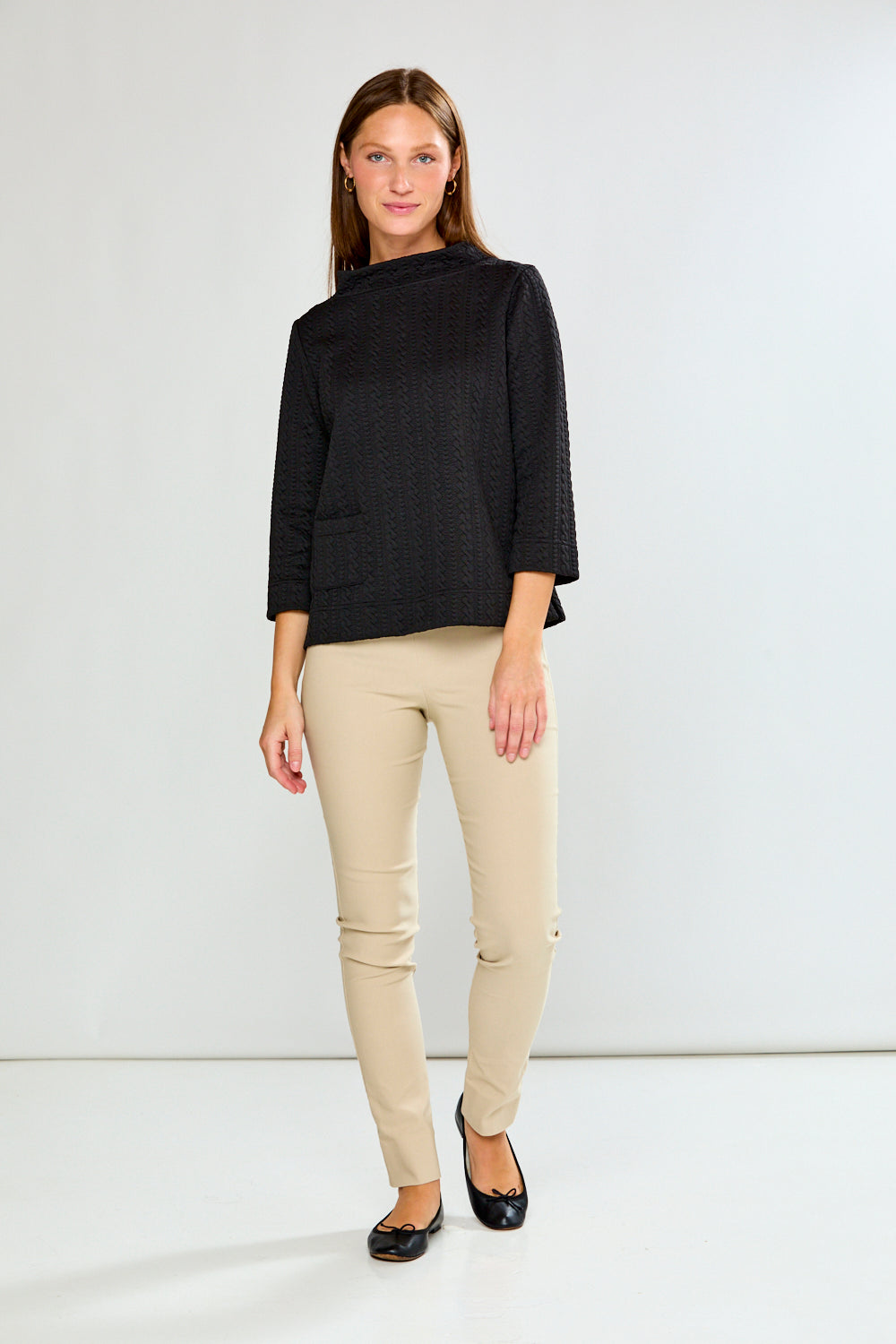 Woman wearing a black textured top and beige pants against a white background