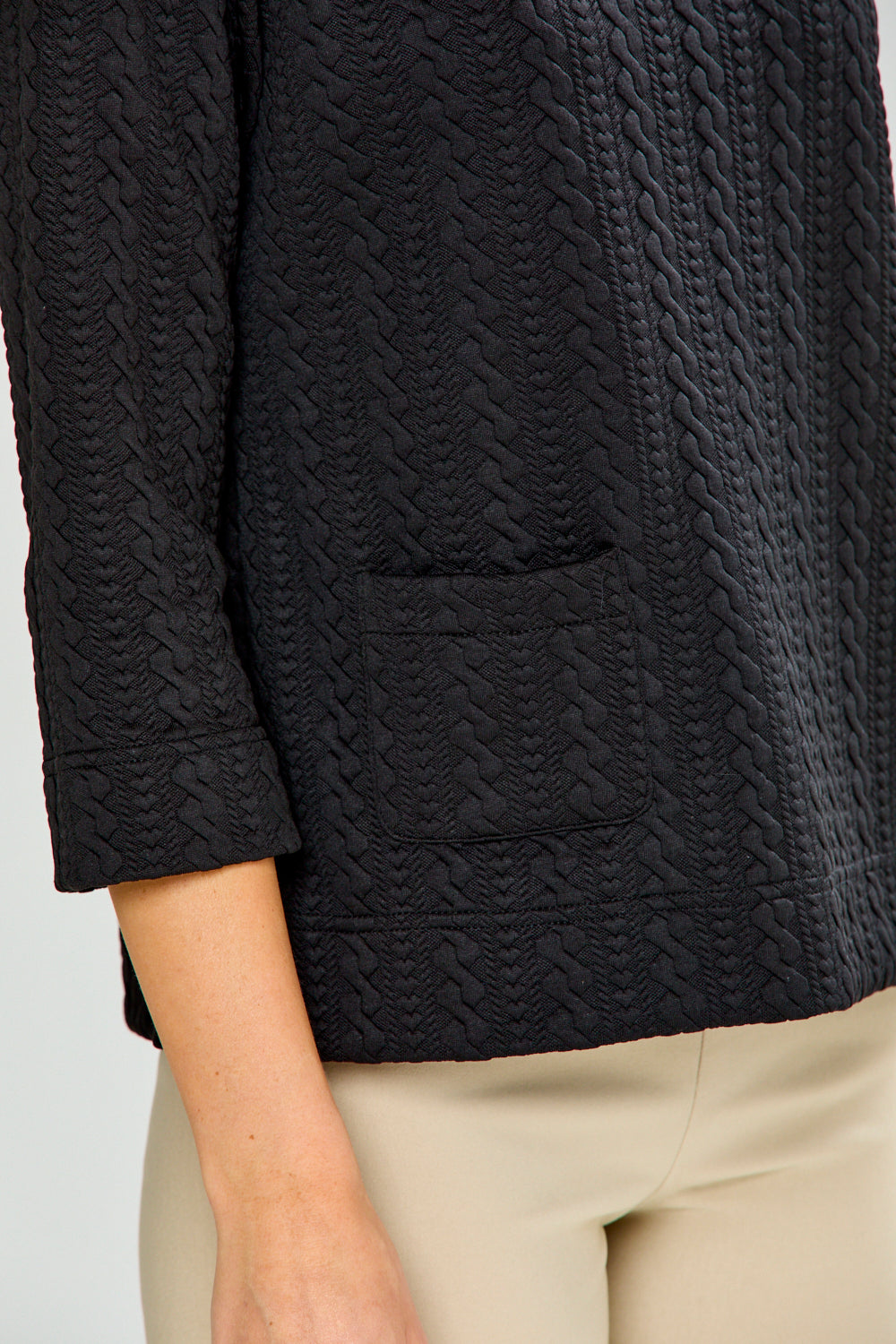 Woman wearing a black textured top and beige pants against a white background