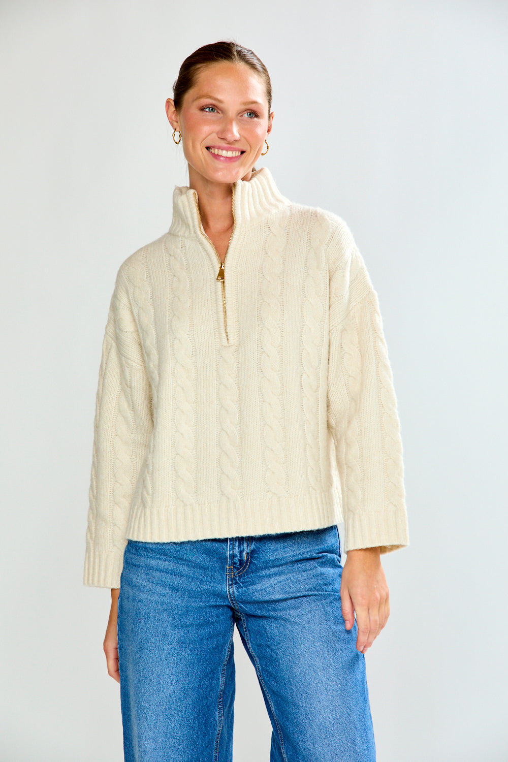 Woman wearing a cream cable knit sweater and blue jeans on a white background