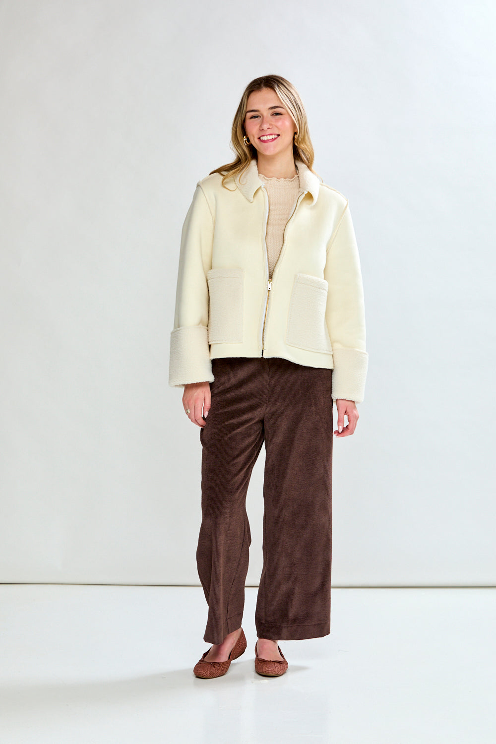 Woman wearing a cream-colored coat over a beige sweater and brown pants on a white background