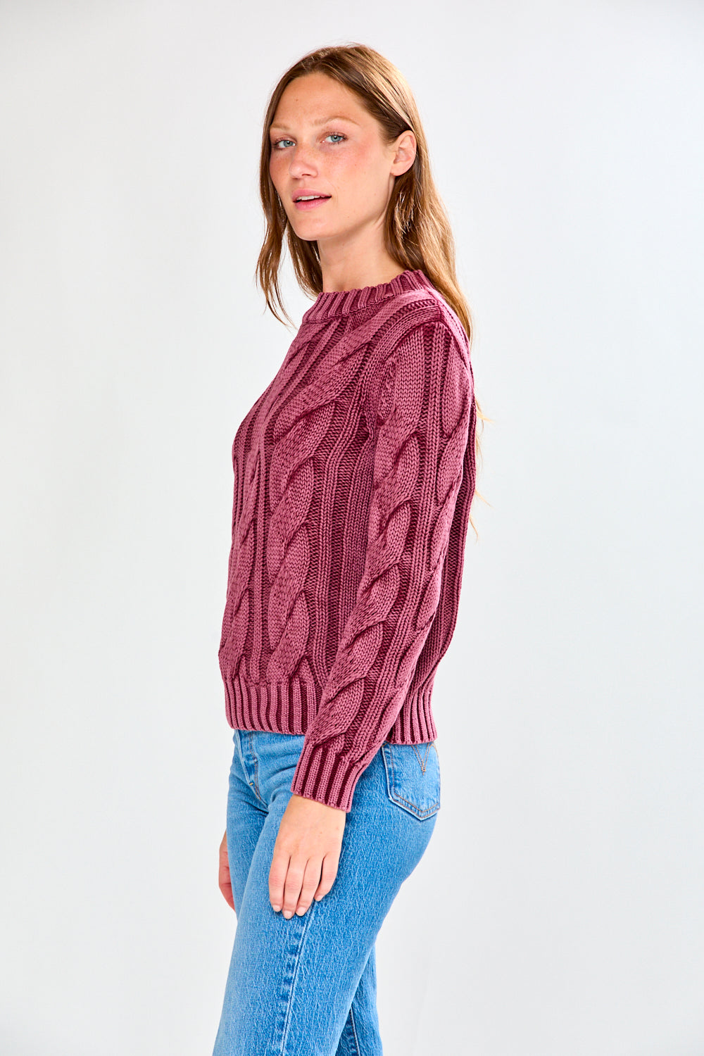 Woman wearing a raisin cable knit sweater and blue jeans on a white background