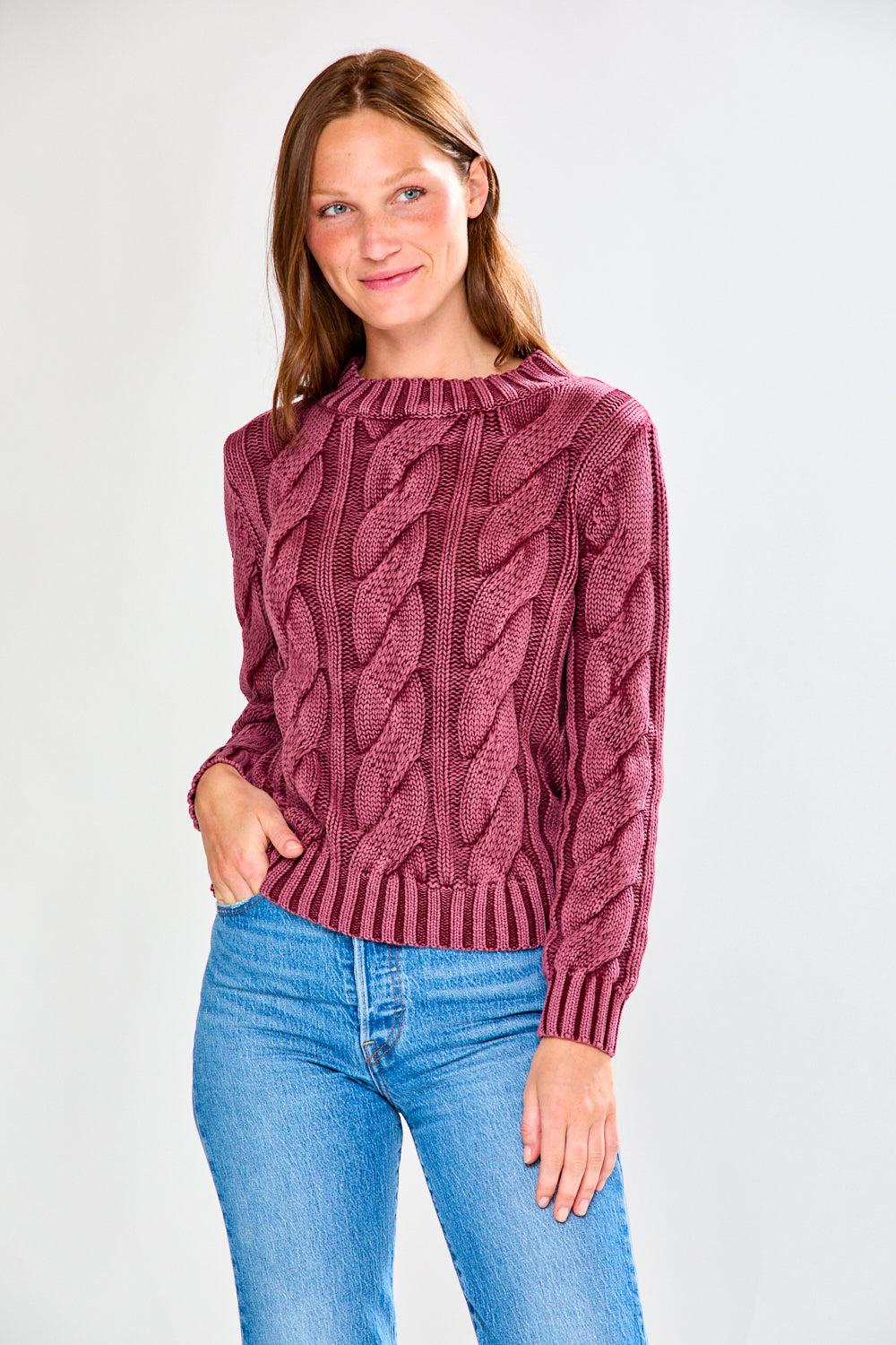 Woman wearing a raisin cable knit sweater and blue jeans on a white background