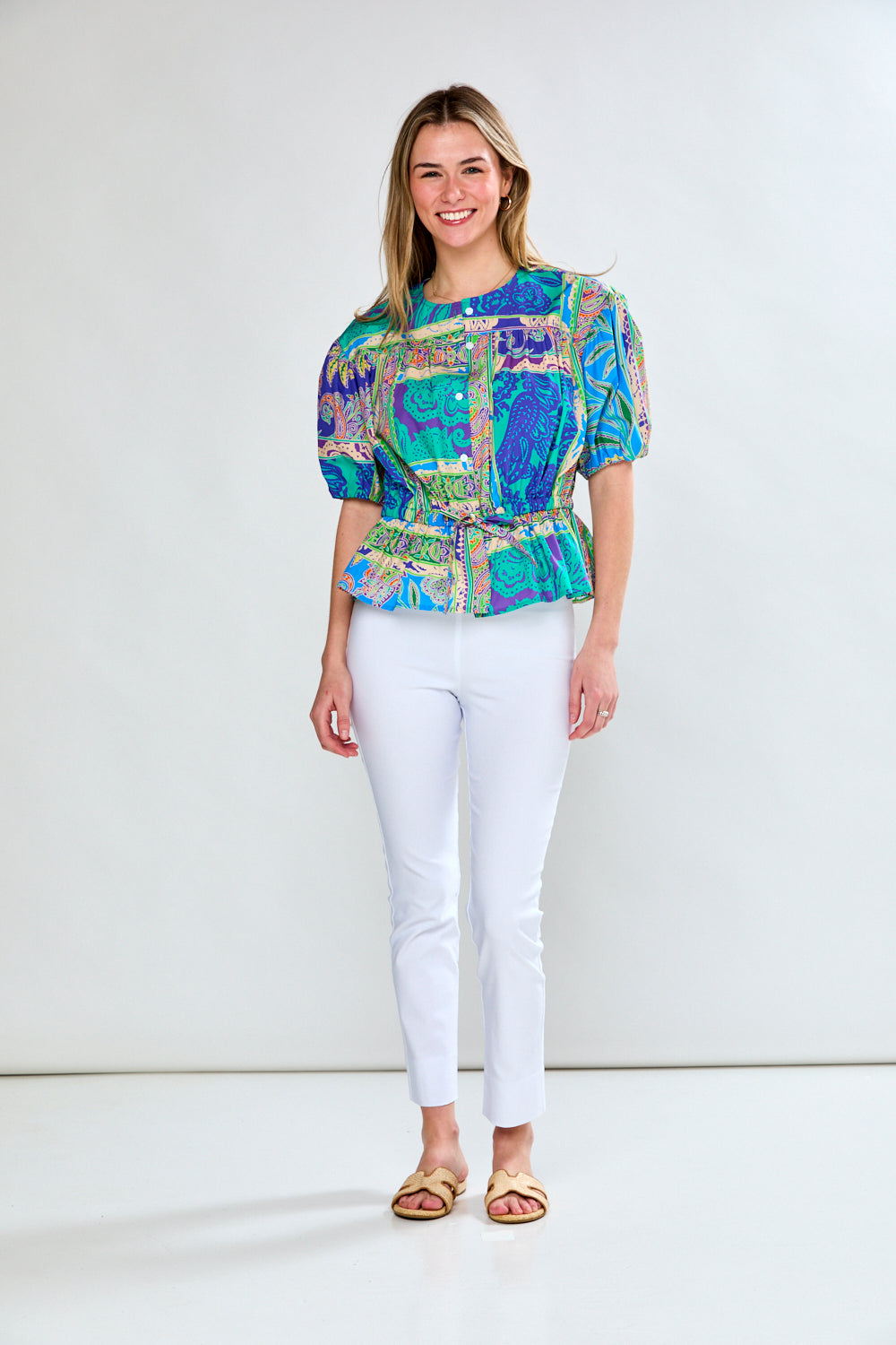A woman wearing a multicolored paisley tiles print blouse with a jewel neckline and puff sleeves, paired with white pants.