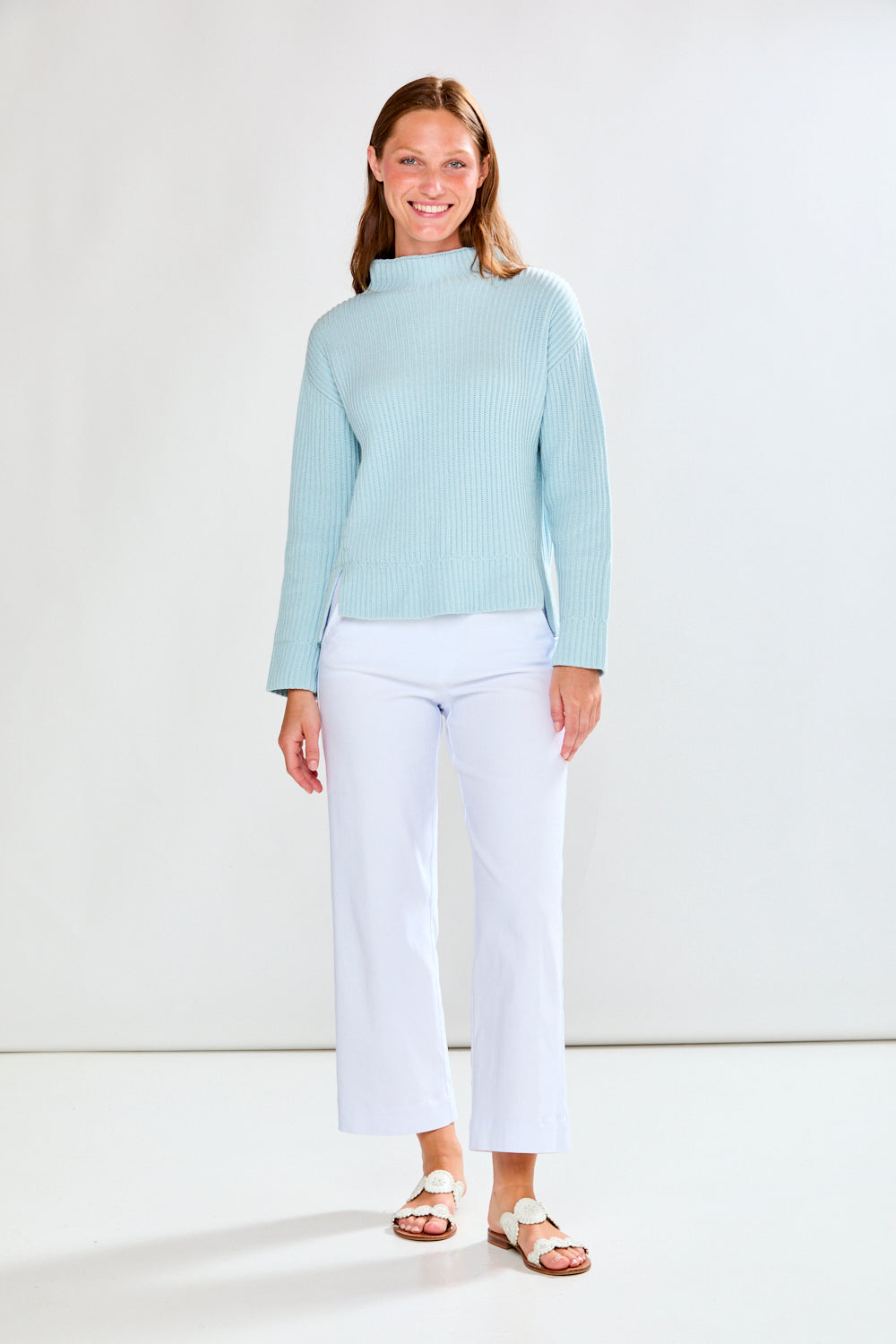 Woman wearing a light blue ribbed sweater and white pants on a plain background