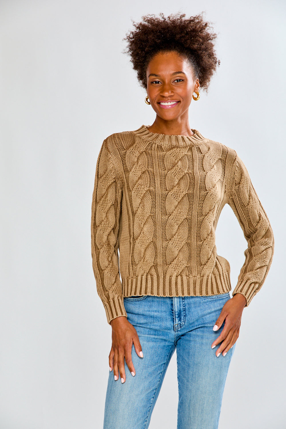 Woman wearing a toffee cable knit sweater and blue jeans on a white background