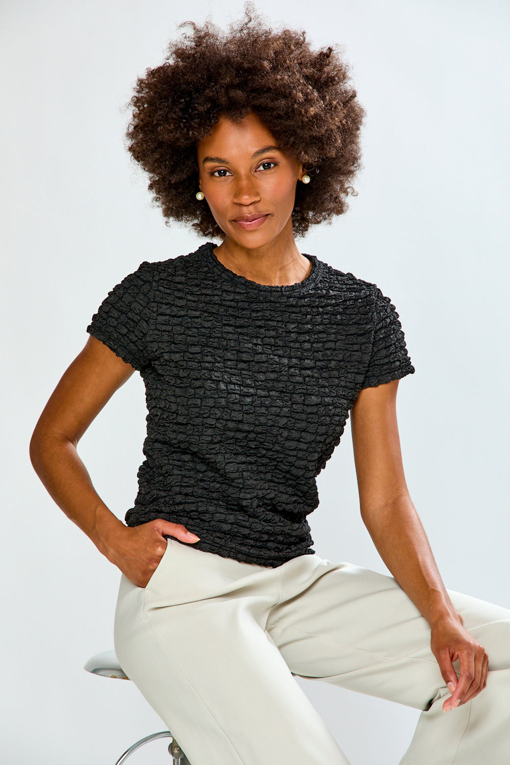 Woman wearing a textured black top and light-colored pants against a plain background
