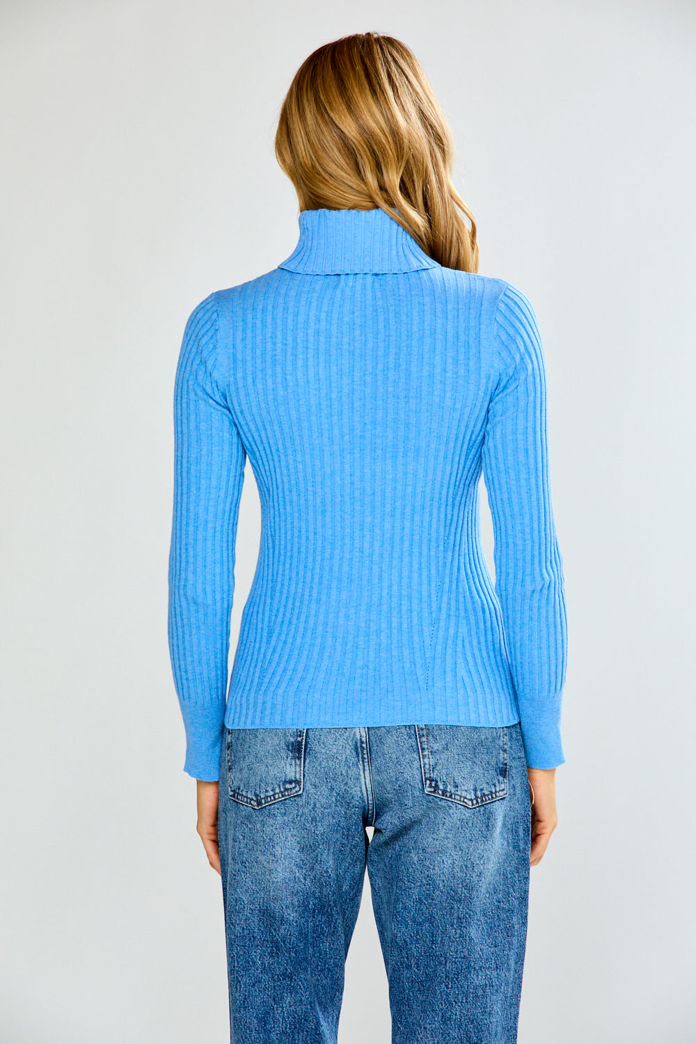 Woman wearing a blue ribbed turtleneck sweater and jeans on a plain background