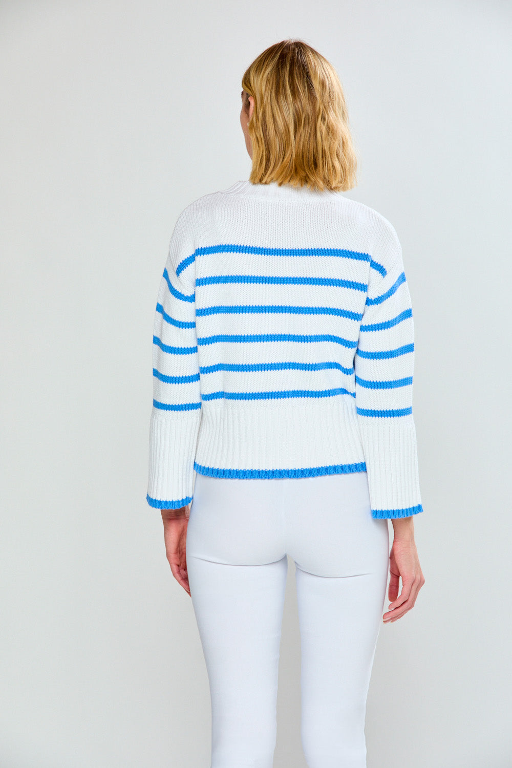 Person wearing a white sweater with blue stripes and white pants on a plain background