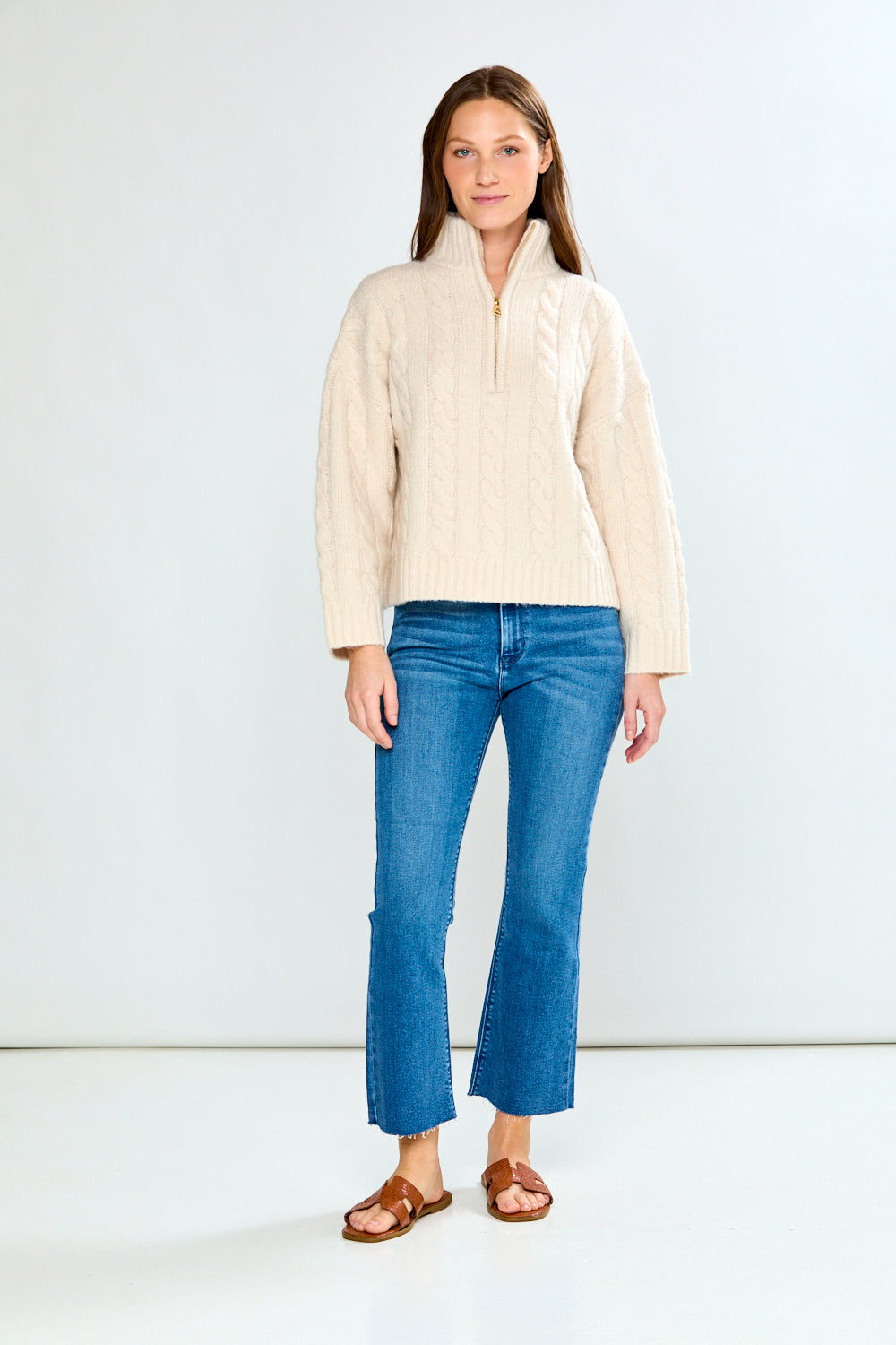 Woman wearing a oatmeal cable knit sweater and blue jeans on a white background
