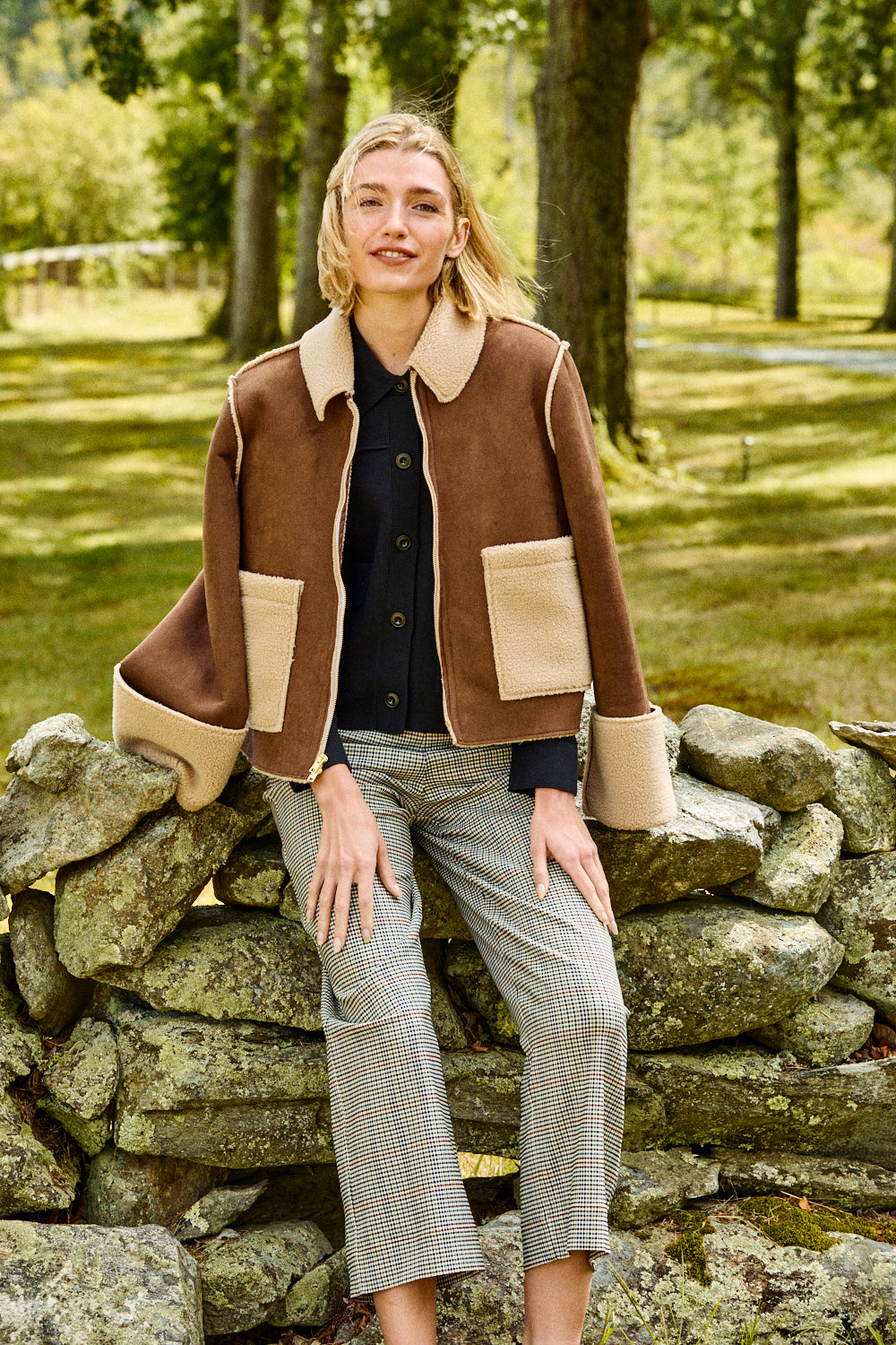 Woman in a brown and beige jacket and plaid pants sitting on a stone wall in a park.