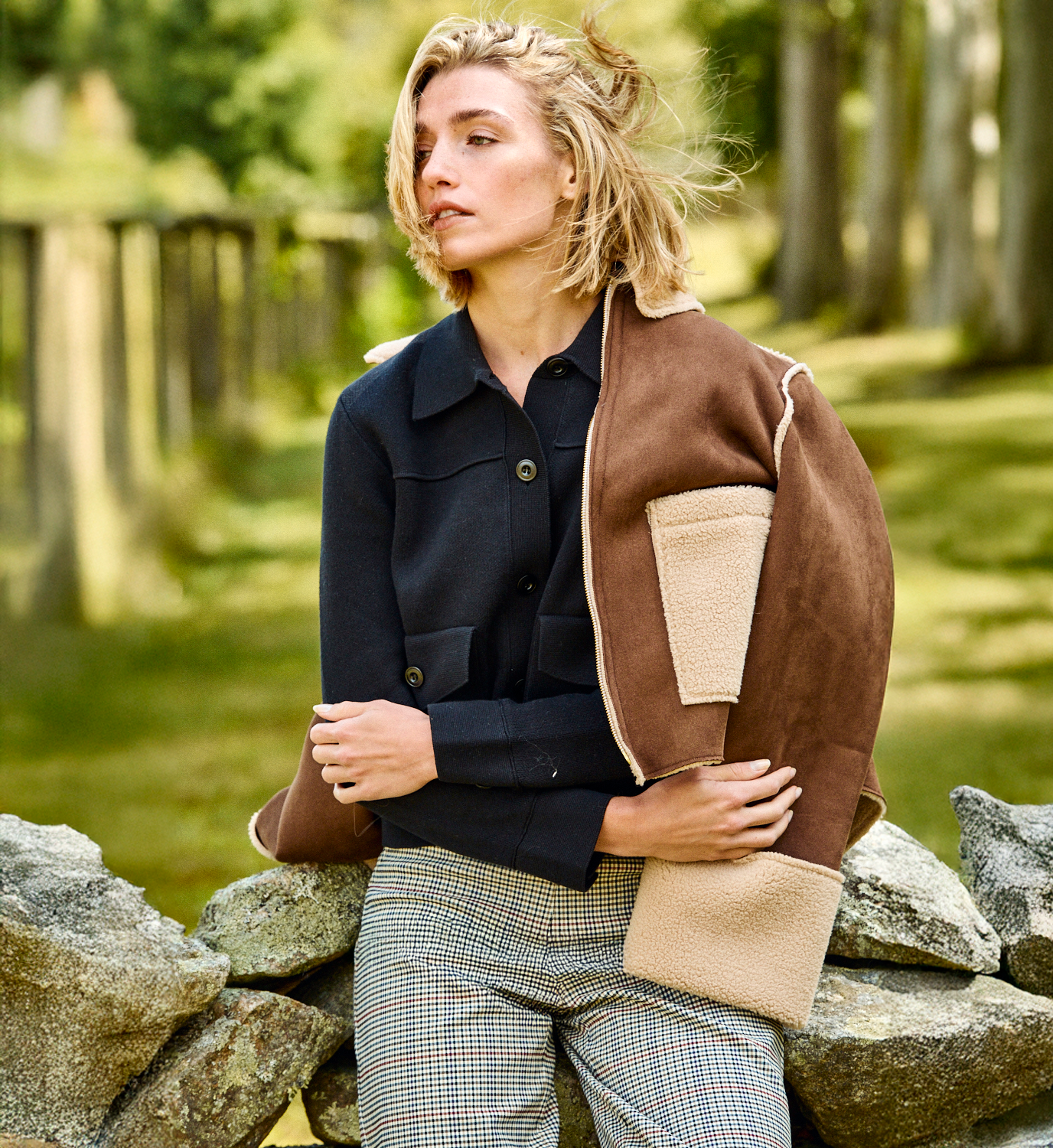 A woman sitting on a rock wall in a field wearing a sweater, pants, and a jacket draped over her shoulders.