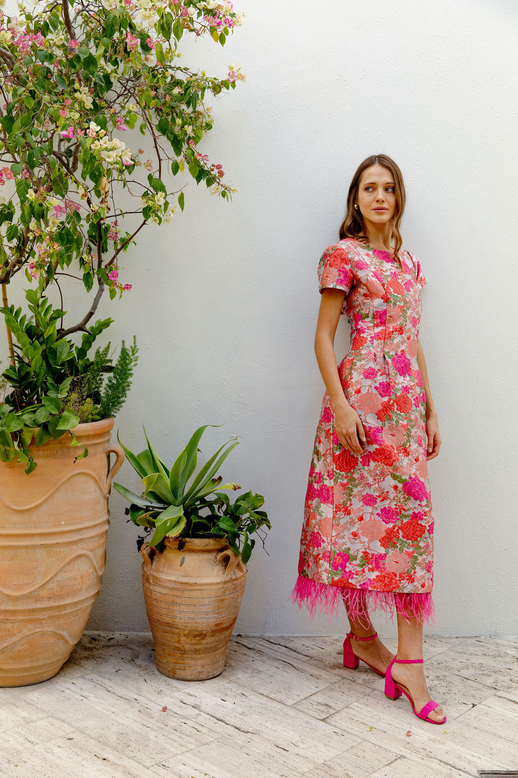 Woman in a floral dress standing next to potted plants against a white wall.