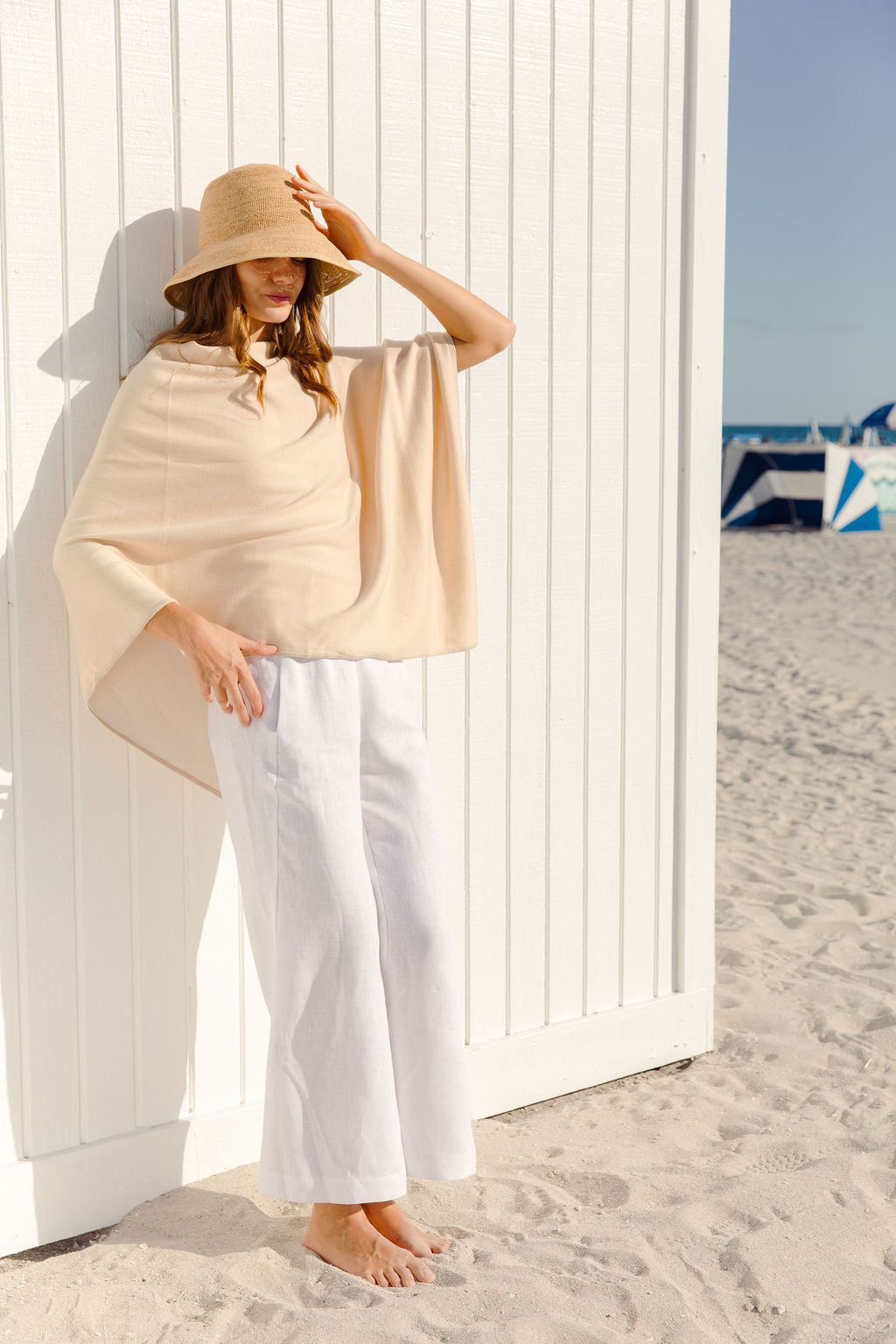 Woman in a beige poncho and white pants standing on a sandy beach with a white wall and blue and white striped towel in the background.