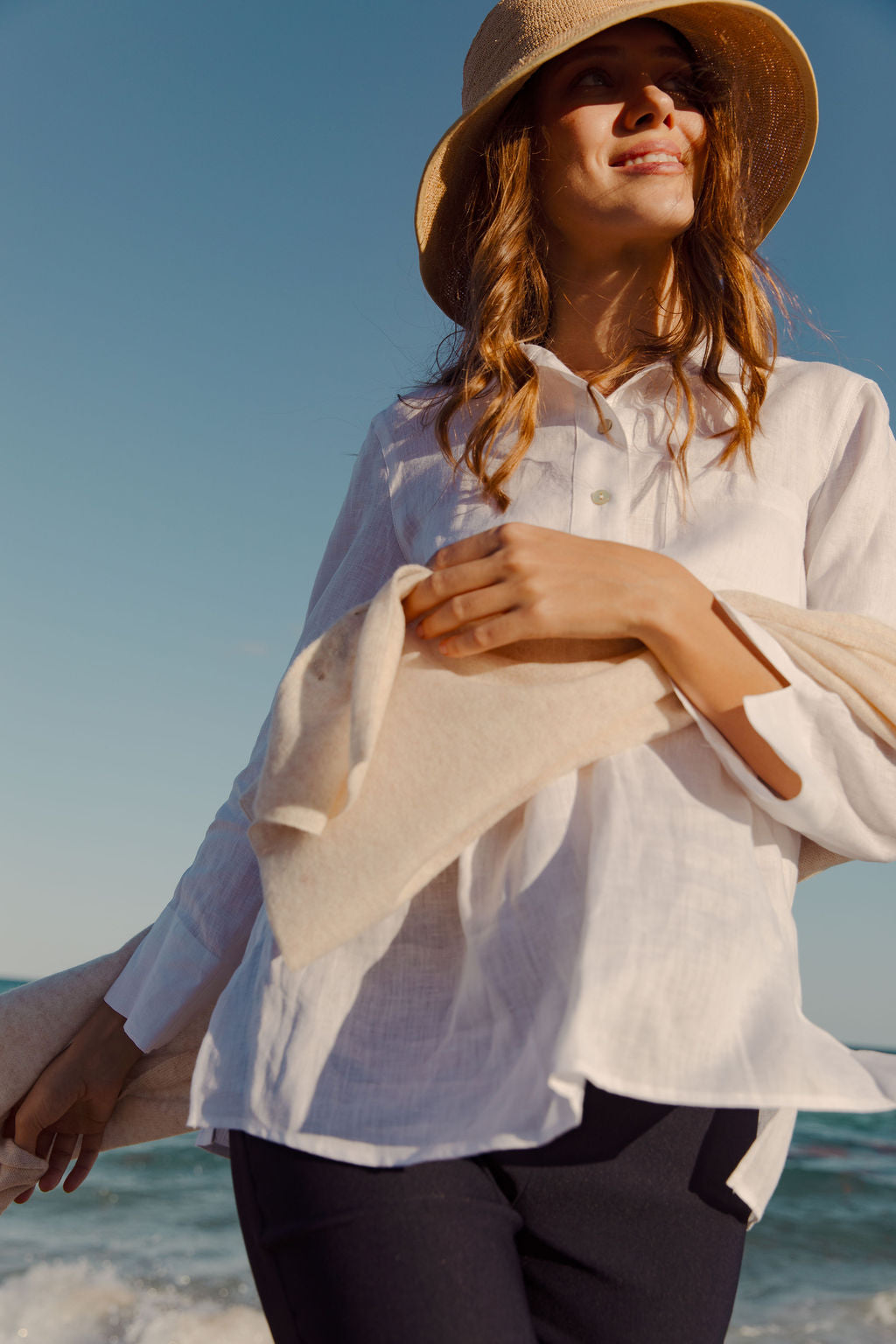 Woman on a beach wearing a white shirt and straw hat with a clear blue sky.