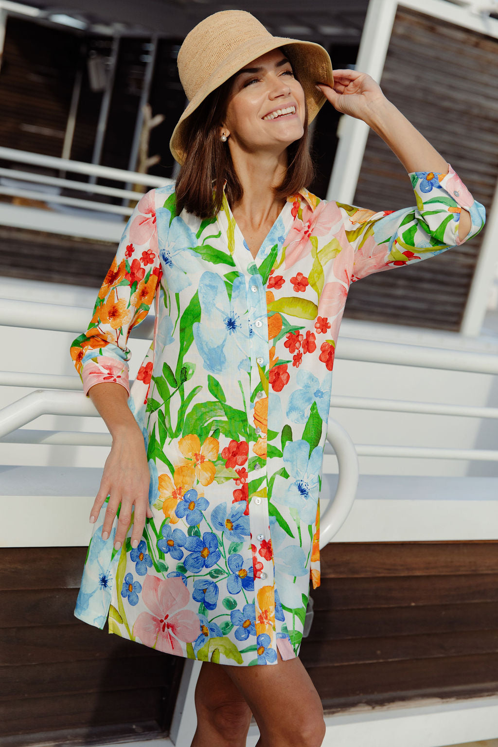 Woman wearing a colorful floral dress and hat on a wooden deck.