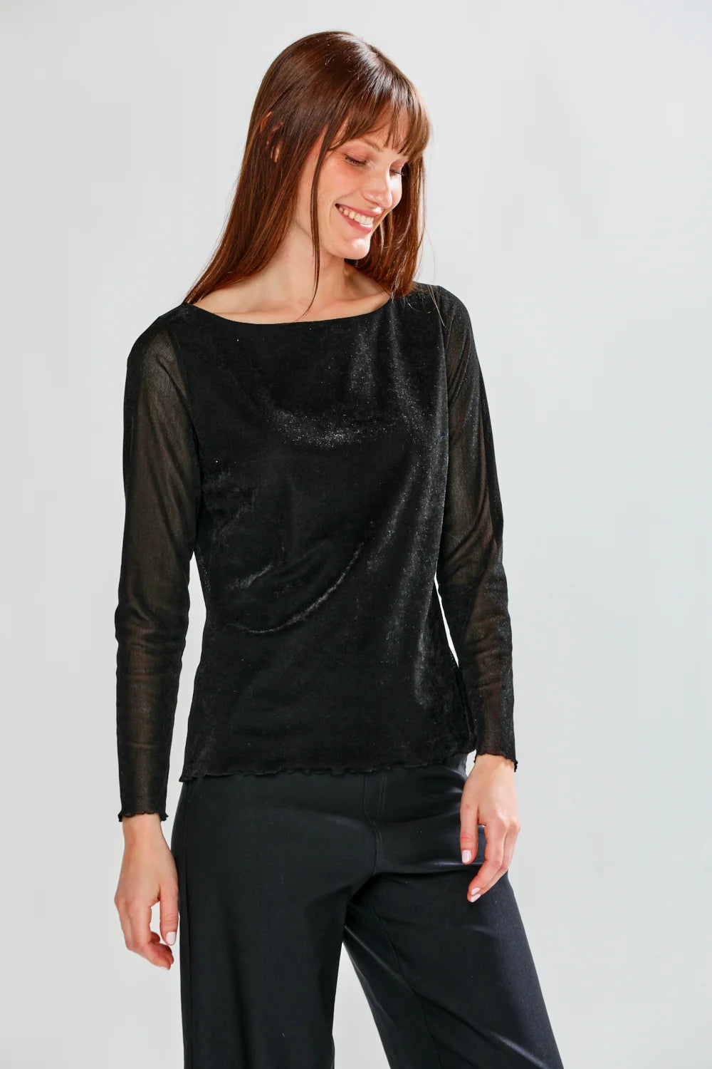 The Selena Top in Velvet - Black worn by a smiling model with sheer long sleeves and a boat neckline in a chic black velvet knit top