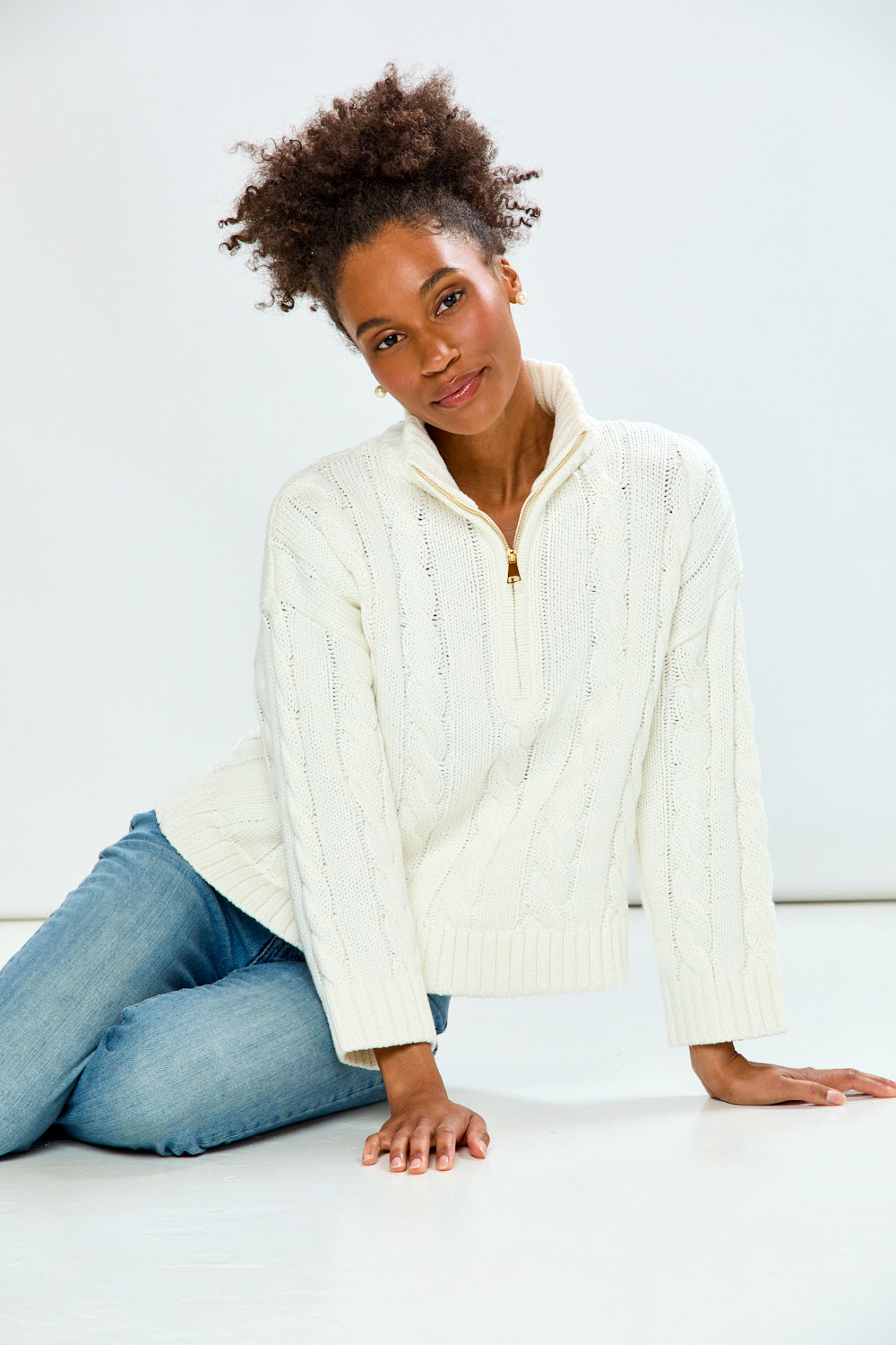 Woman wearing a white cable knit sweater and blue jeans sitting on a white surface.