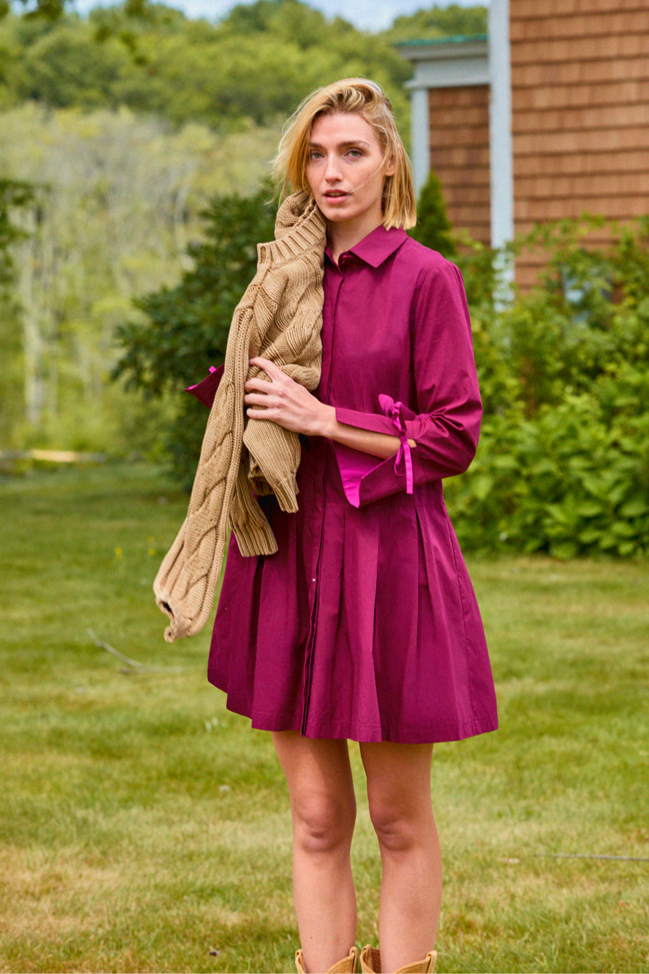 Woman in a purple dress holding a beige jacket outdoors with greenery and a wooden building in the background