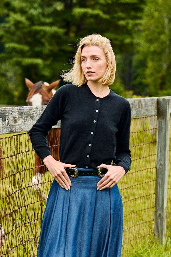 Woman in a black cardigan and blue skirt standing next to a wooden fence with a horse in the background.
