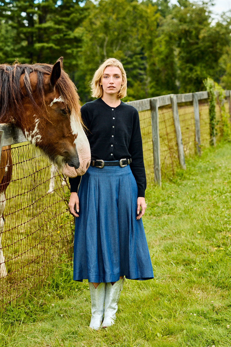 Woman in a blue skirt standing next to a horse in a grassy field with a wooden fence and trees in the background.