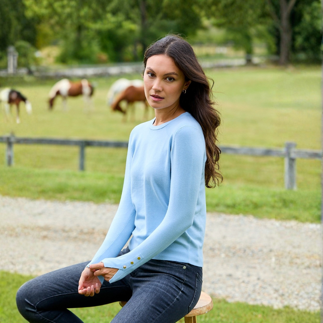 A woman sitting on a wooden stool in a grassy field, wearing a light blue long-sleeve knit sweater with gold button details on the cuff and dark jeans. Horses are visible in the background.