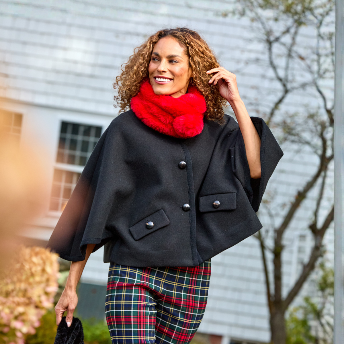 A woman wearing a black cape-style coat with silver buttons, a bright red fuzzy pom-pom scarf, and red, green, and blue plaid pants, standing outdoors in front of a house.