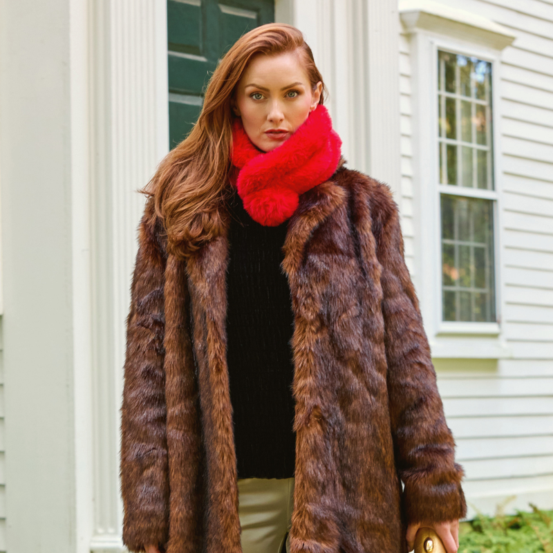 A woman wearing a brown faux fur coat, black knit sweater, and a bright red fuzzy scarf, standing in front of a white house.