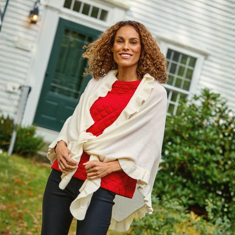 A smiling woman with curly brown hair stands outdoors in front of a white house with a green door, wearing a cream ruffled shawl over a red top and dark pants.