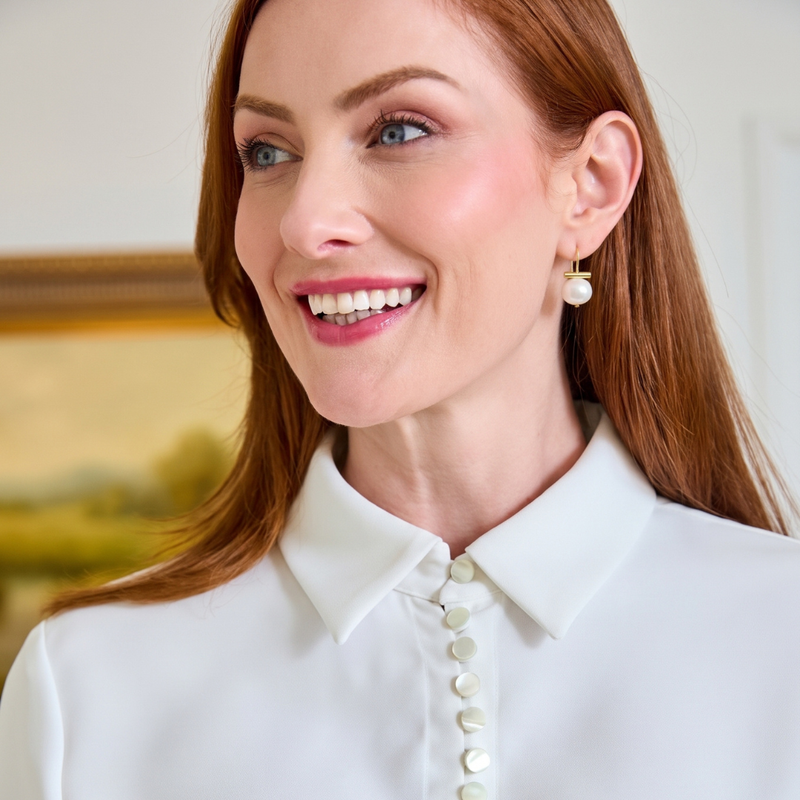 A smiling woman with red hair wearing a white collared blouse with pearl buttons and a pearl drop earring, photographed indoors against a softly blurred background.