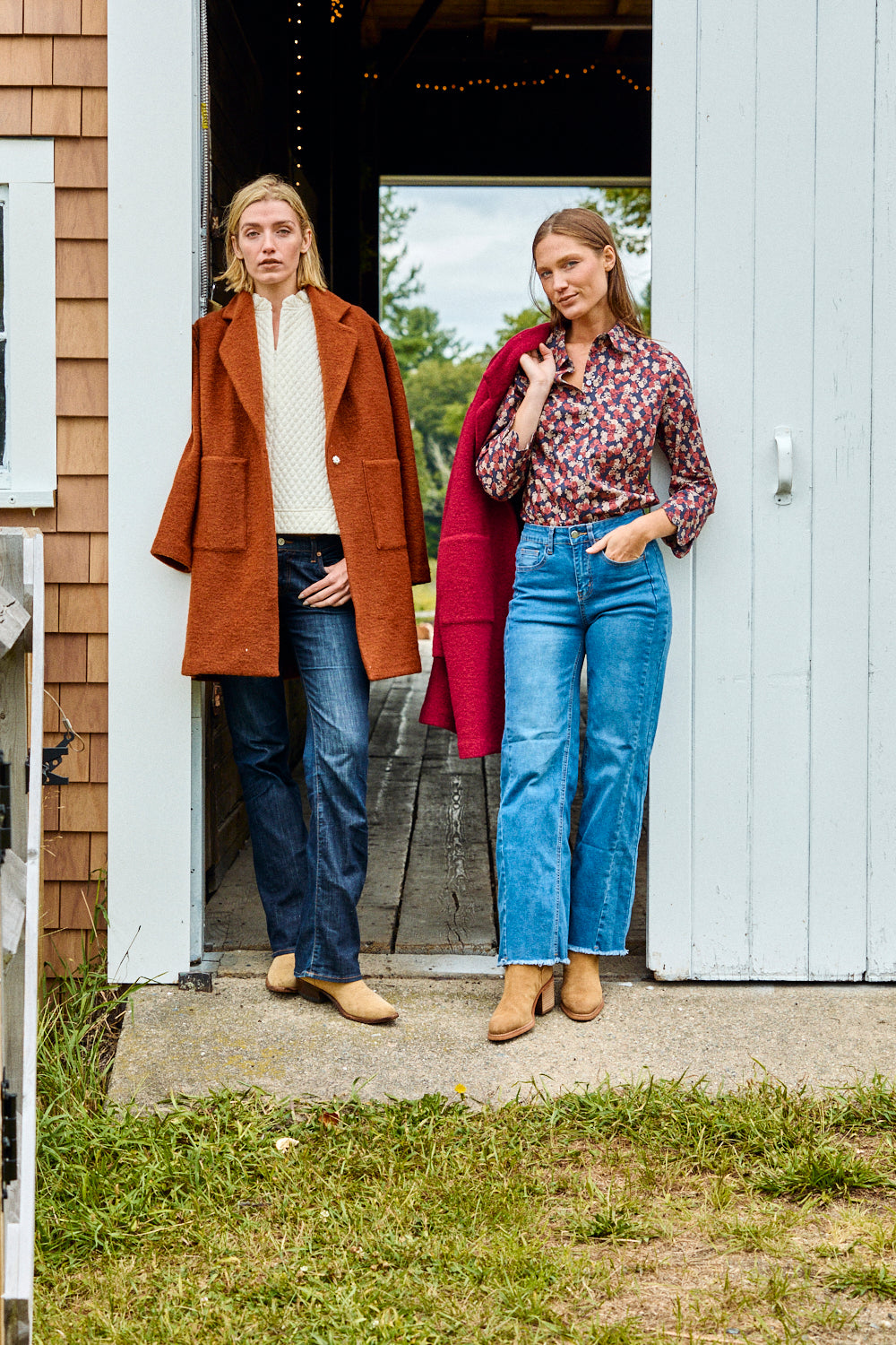 Two women standing outside a building, one wearing a brown coat and blue jeans, the other in a red coat and patterned shirt with blue jeans.