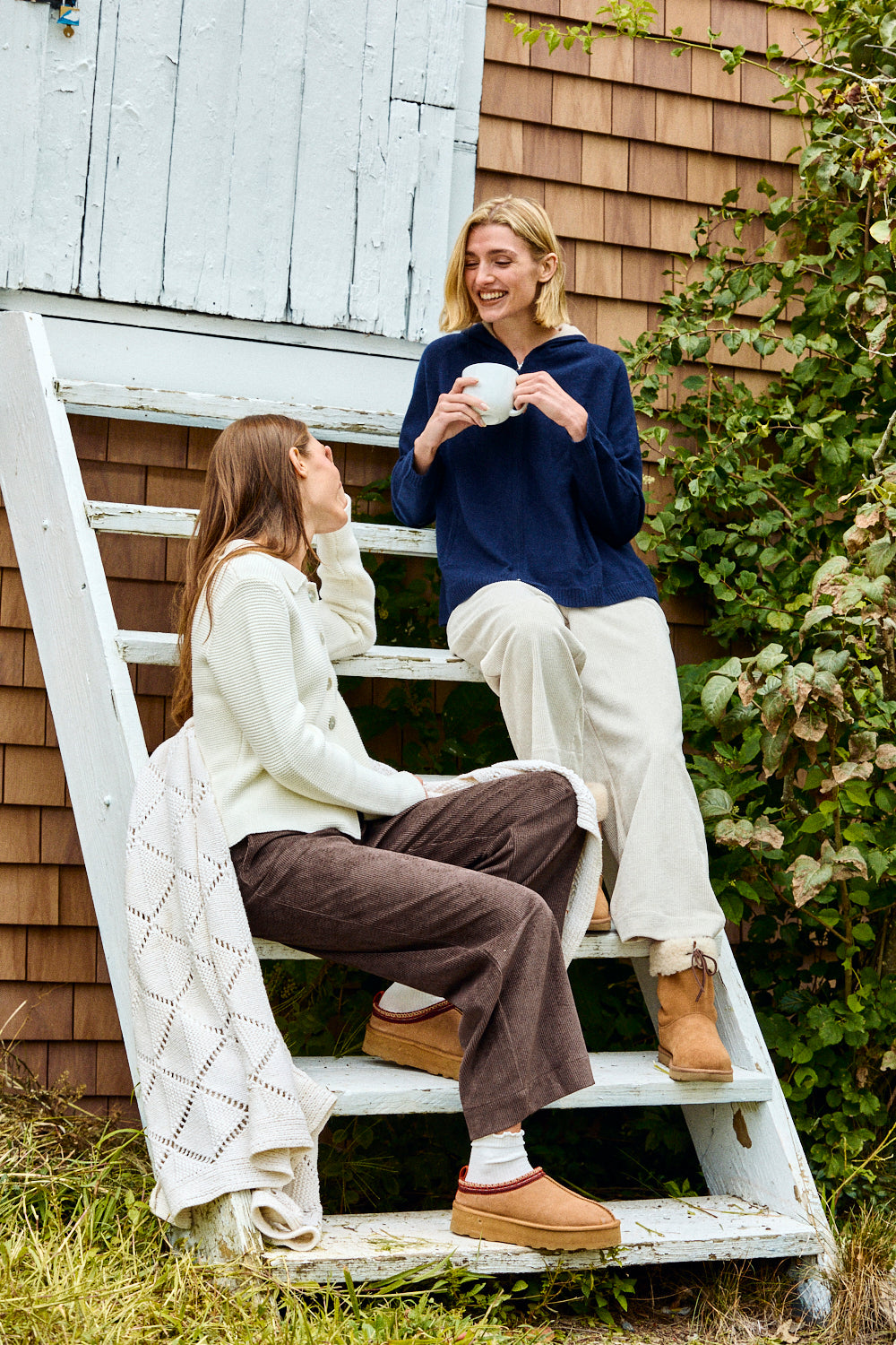 Two women sitting on a white ladder outdoors, one holding a cup.