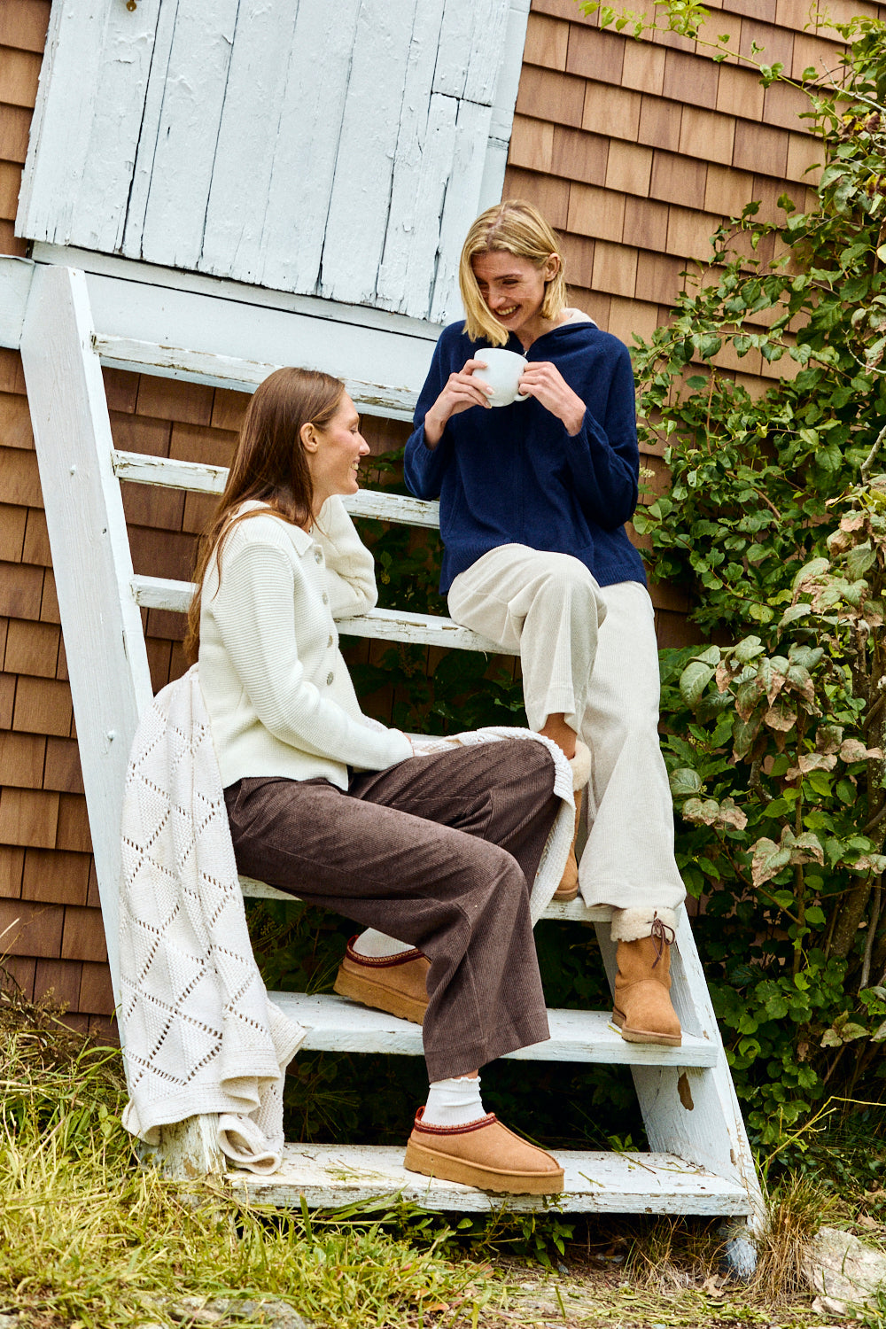 Two women sitting on a white ladder outside a wooden building.