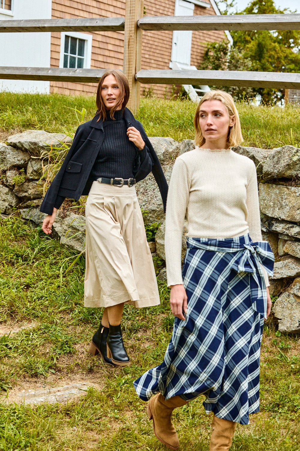 Two women standing outdoors with a stone wall and wooden staircase in the background.