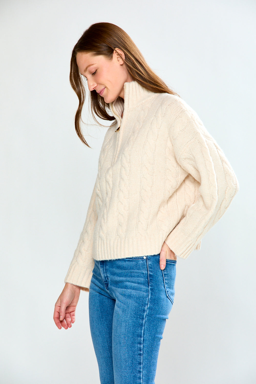 Woman wearing a oatmeal cable knit sweater and blue jeans on a white background