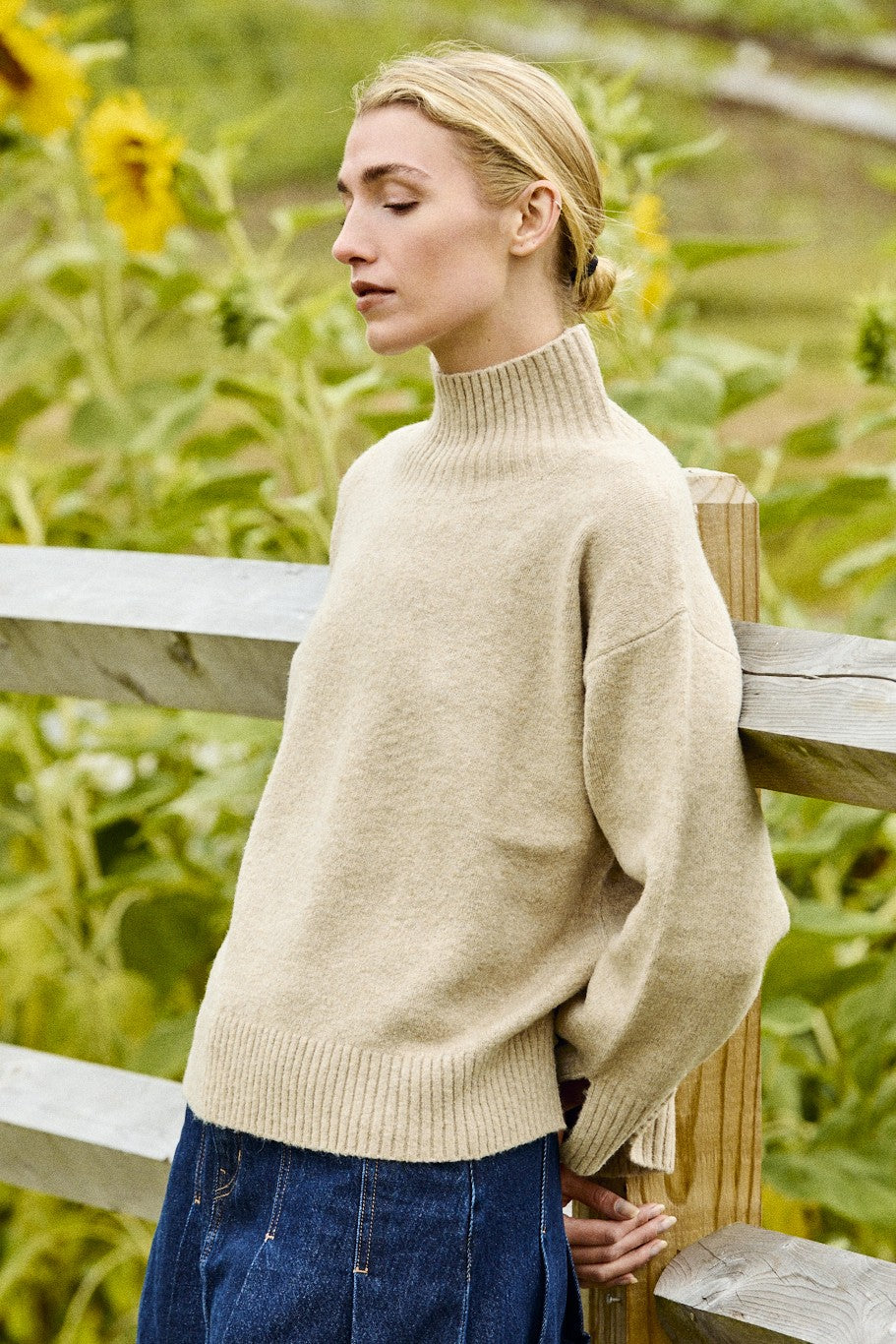 Woman wearing a beige sweater and blue jeans standing in a sunflower field.