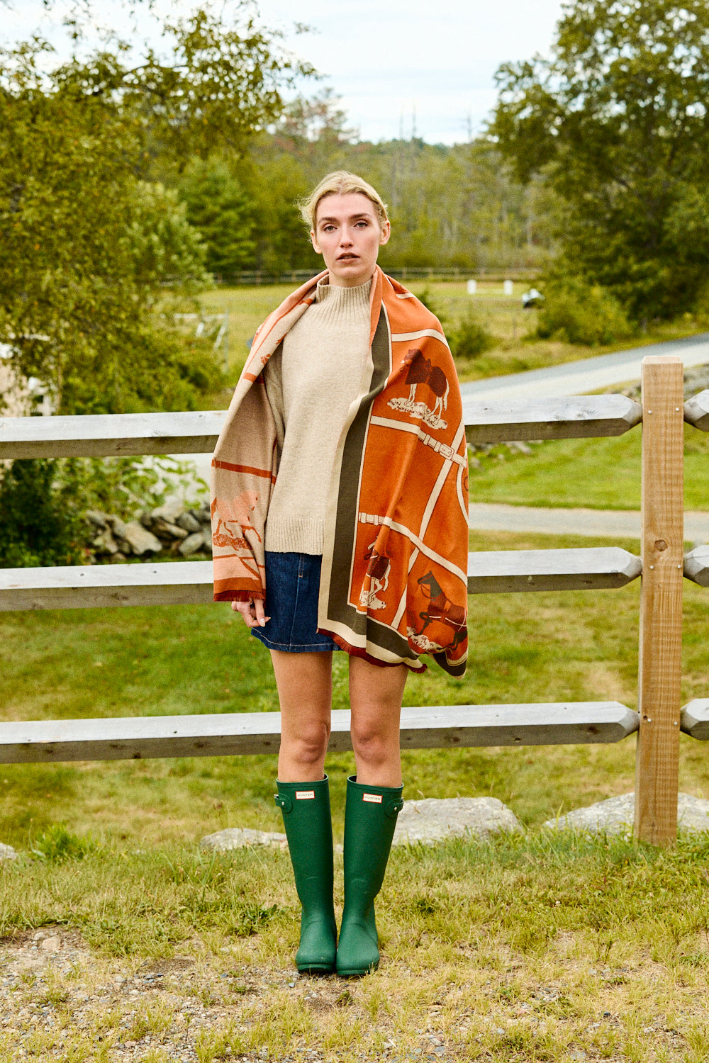 Woman standing outdoors wearing a patterned shawl, beige sweater, blue shorts, and green rain boots.