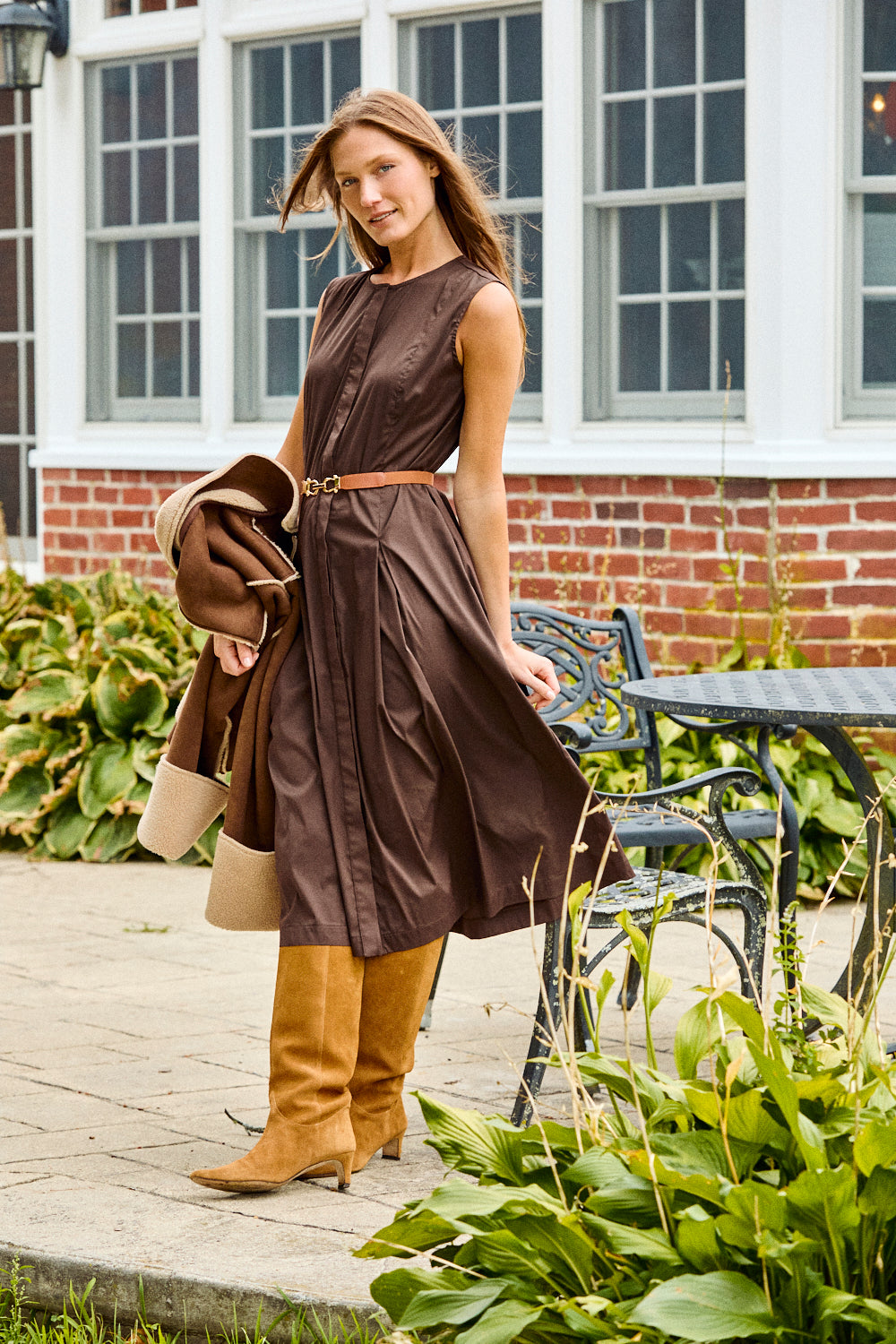 Woman in a brown dress holding a coat in front of a brick building with windows.